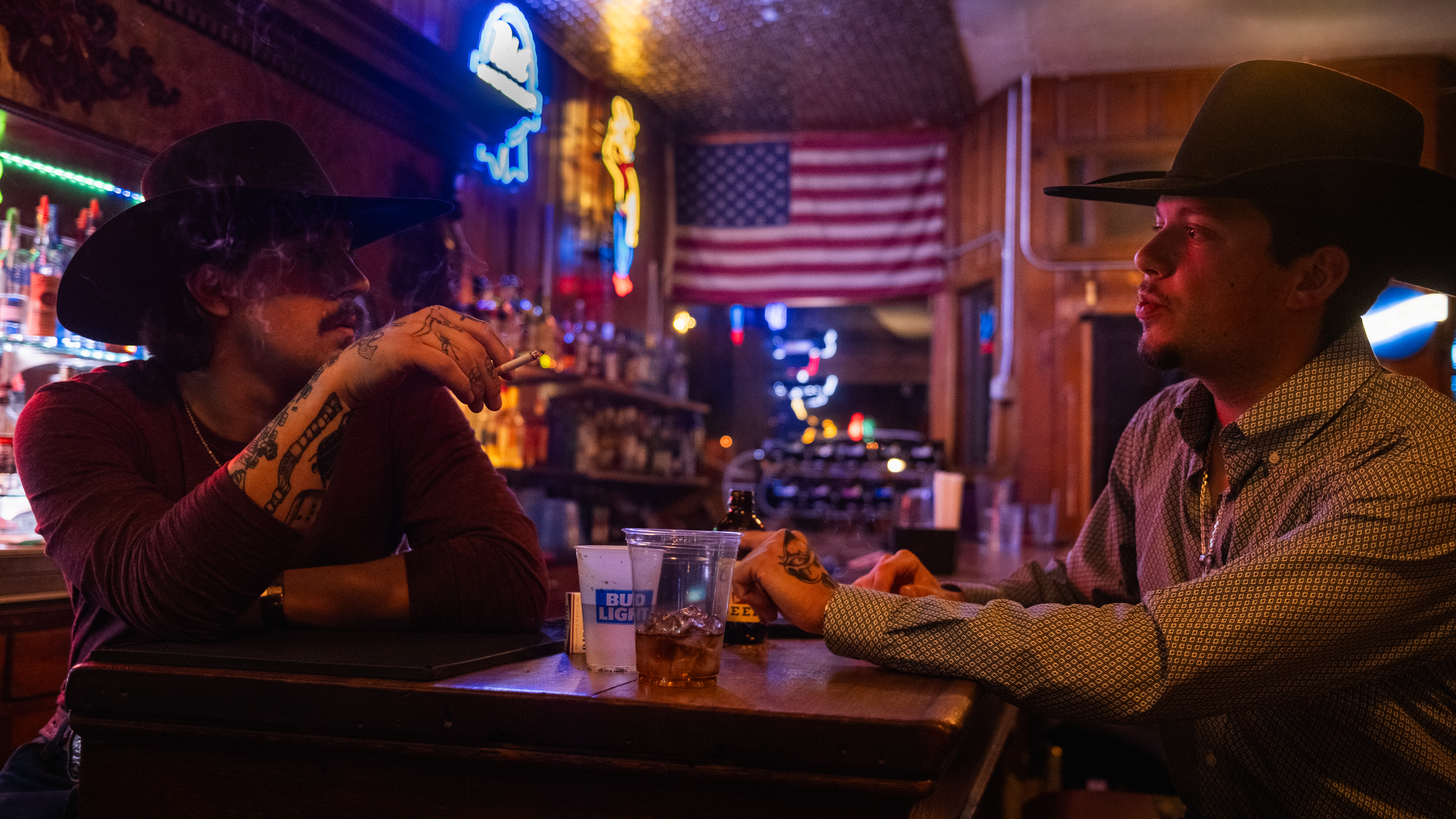 Two men in cowboy hats talk across a bar at night, neon signs and an American flag behind them