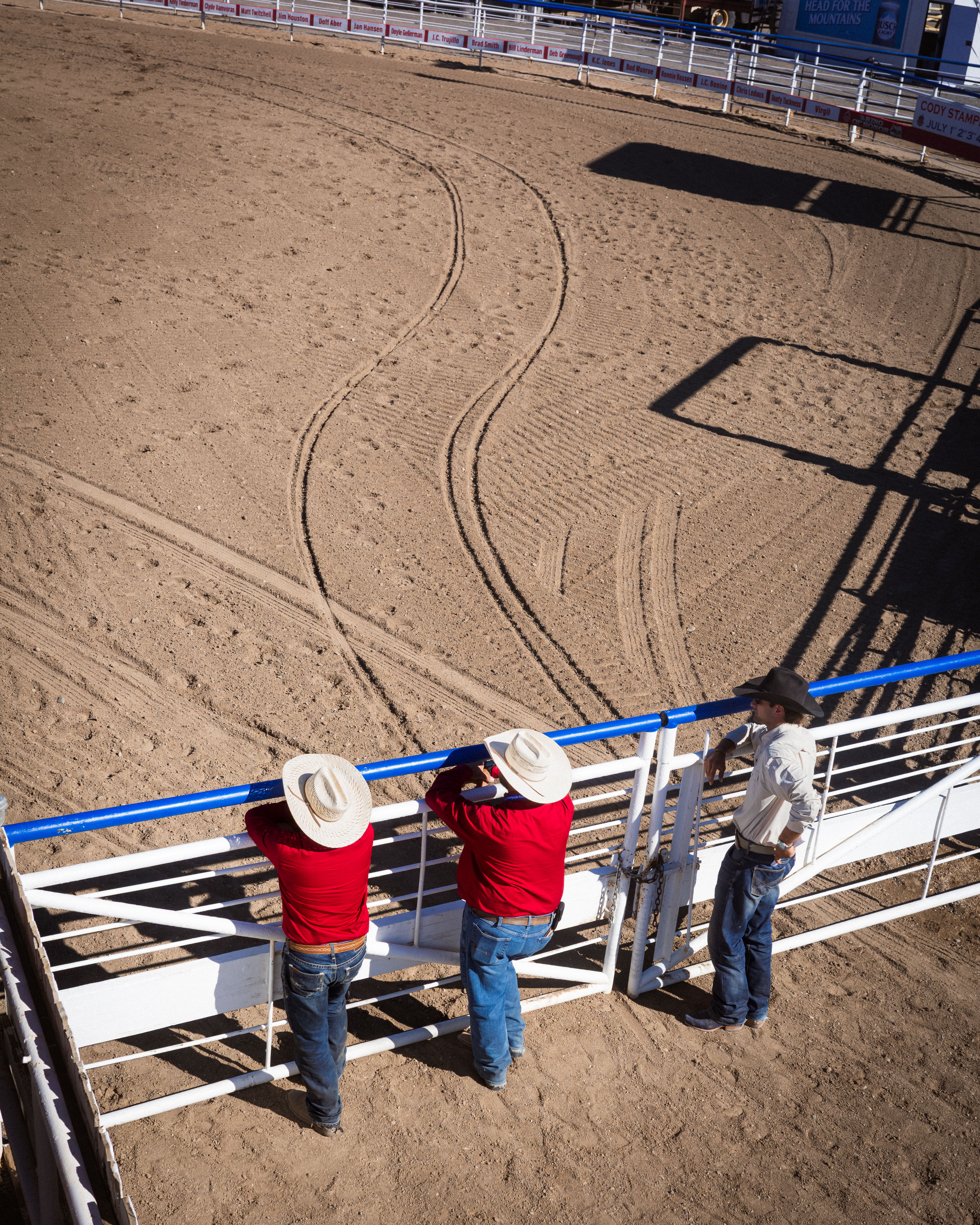 Overhead view of three men in hats leaning on a white arena rail, curved tire tracks in the dirt