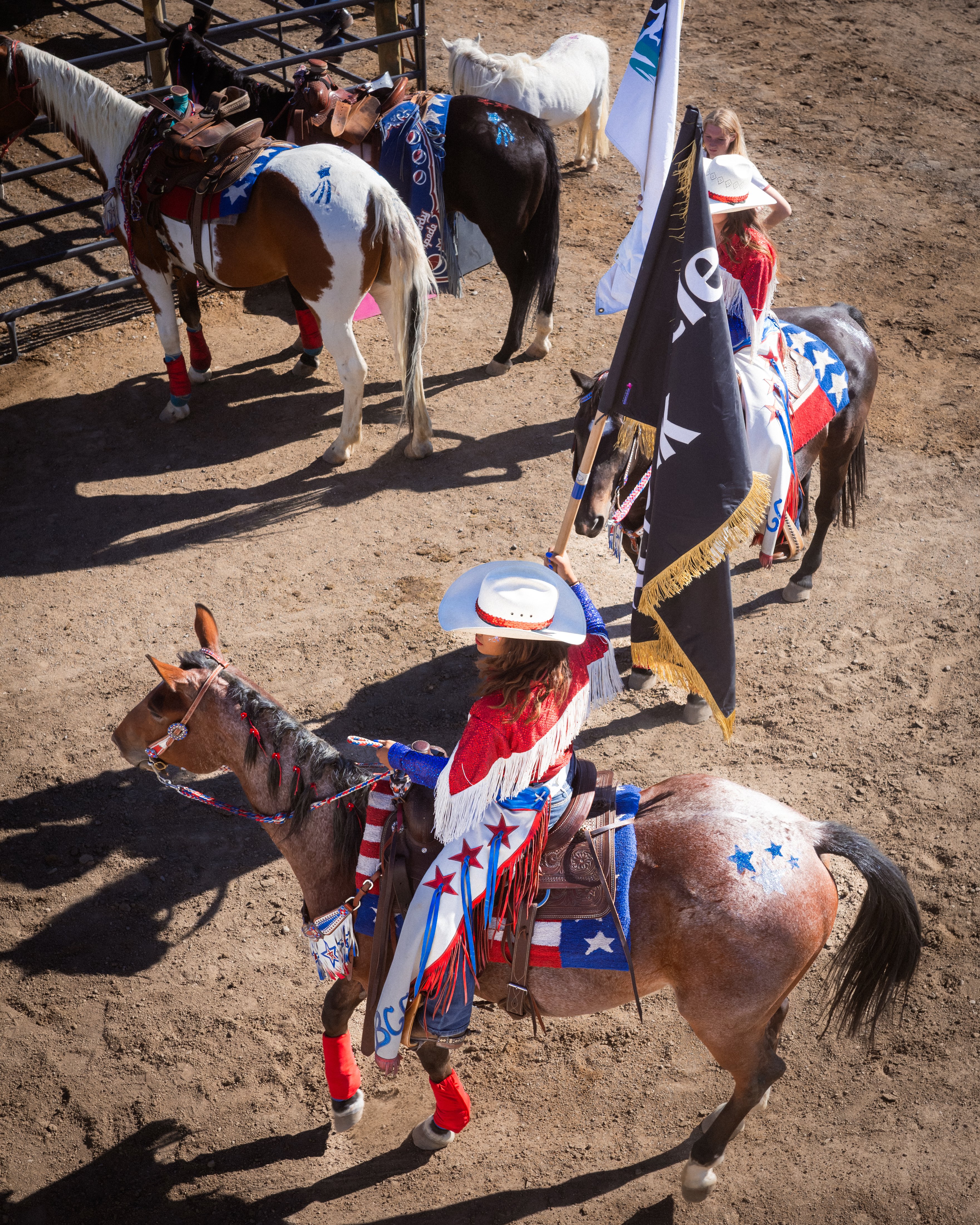 A rodeo queen in red on a spotted Appaloosa carries a team flag across the arena dirt