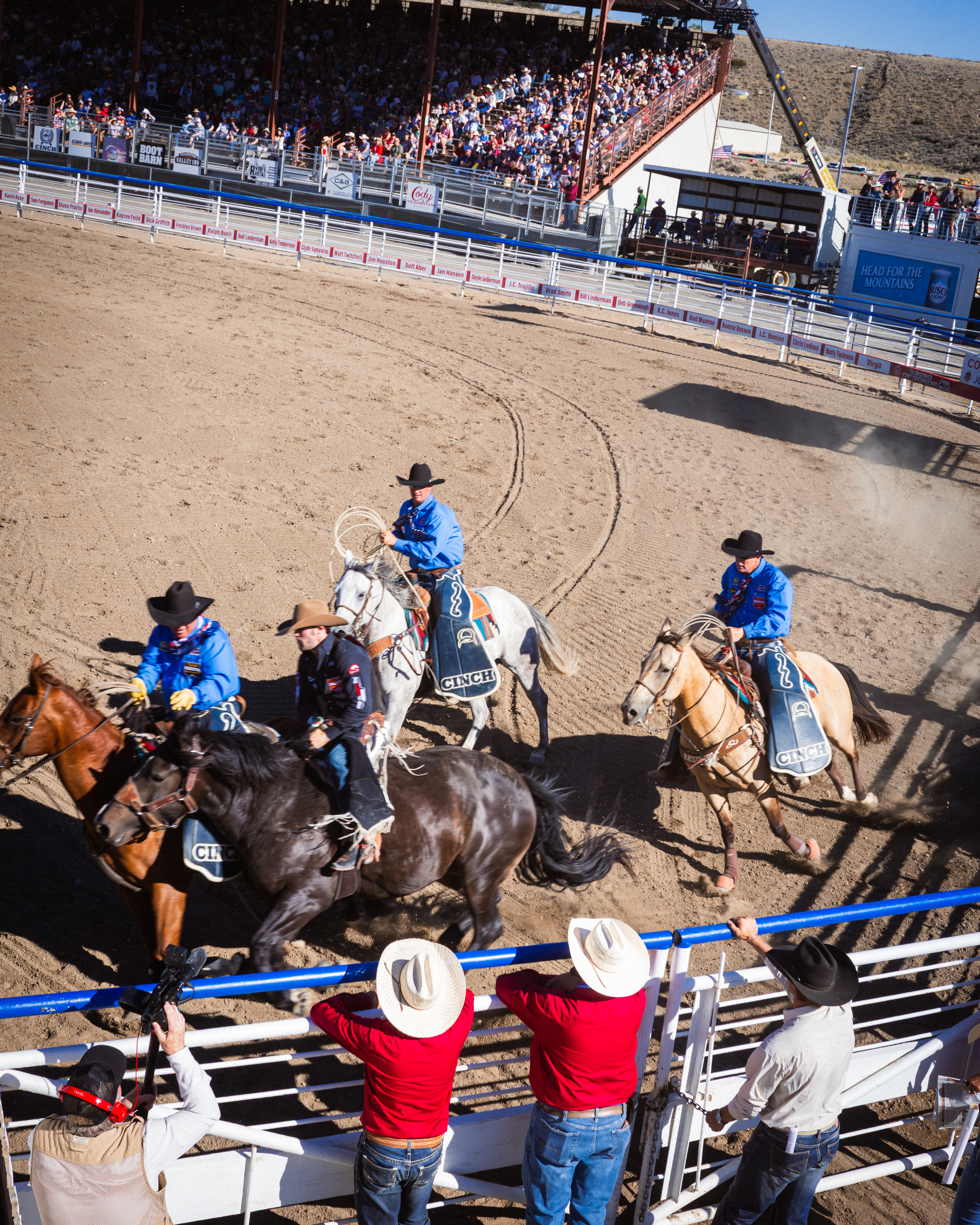 Riders in blue shirts on horseback kick up dust during a rodeo grand entry as spectators watch from the rail