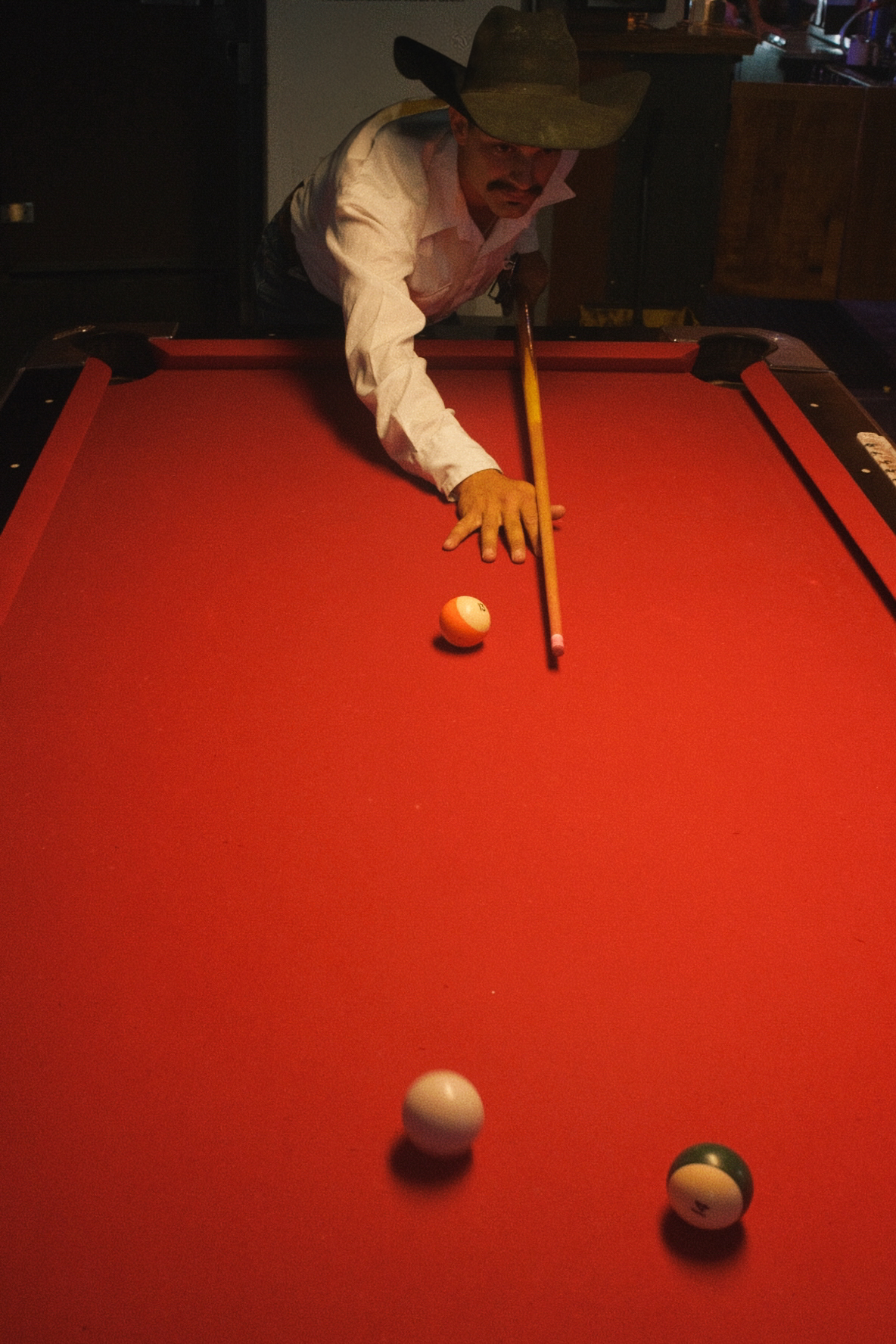 A man in a cowboy hat leans over a red-felt pool table lining up a shot